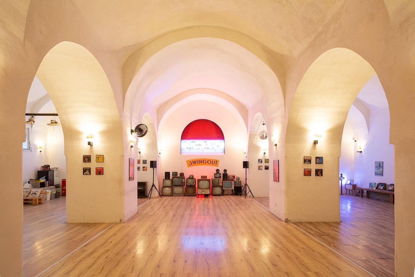 La sala dello Swingout vuota: volte a botte color crema, pavimento in parquet, insegna SWINGOUT e una pila di televisori vintage sul fondo